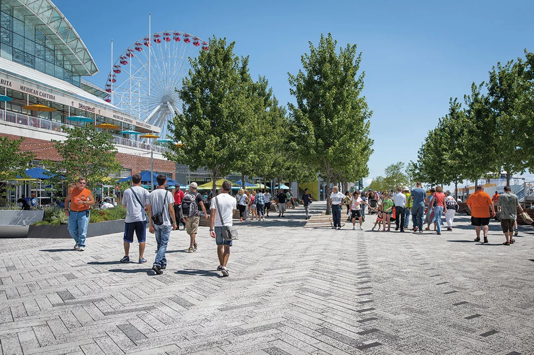 Pedestrians stroll across the Navy Pier on light grey pavers laid in a herringbone pattern.